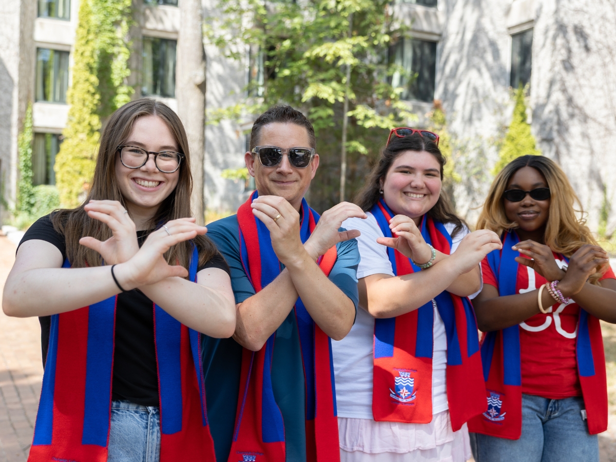 Four people standing outside together holding up their hands to form two letter C's