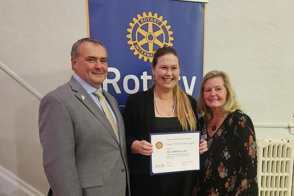 Three people standing infront of a Rotary sign, one is holding a certificate