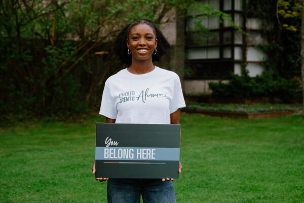 A woman standing outside smiling at the camera, holding a sign that says "You Belong Here"