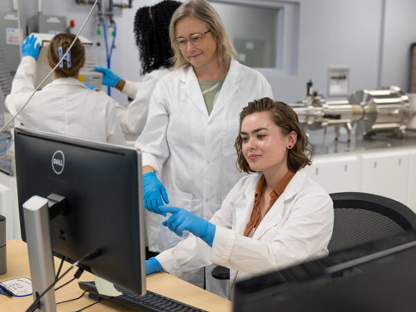 2 women in lab coats and gloves pointing at computer