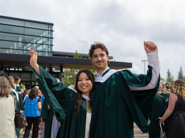 A woman and man wearing convocation regalia with their arms up smiling at the camera