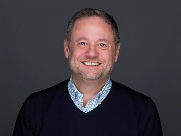 White male with short hair wearing collared shirt under sweater, smiling at camera