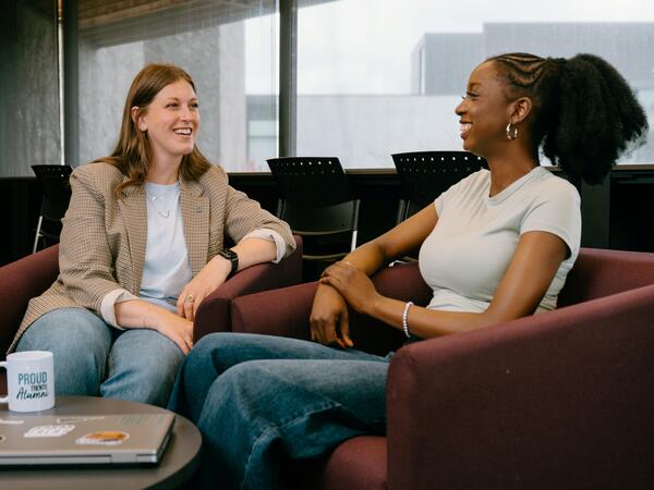 2 women sitting in chairs talking with mug and laptop on coffee table in front of them