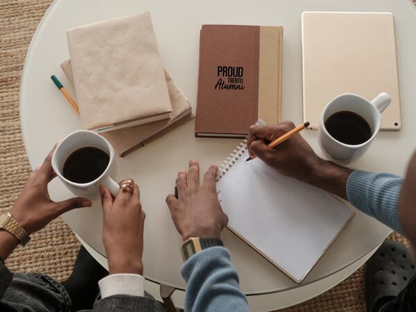 An overhead shot of two people working, one is writing on a pad of paper and the other is drinking coffee