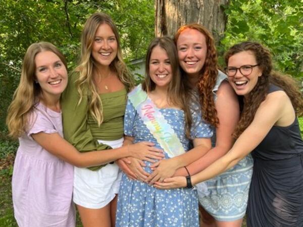 A group of 5 women standing outside with their hands on the pregnant belly of the woman in the middle