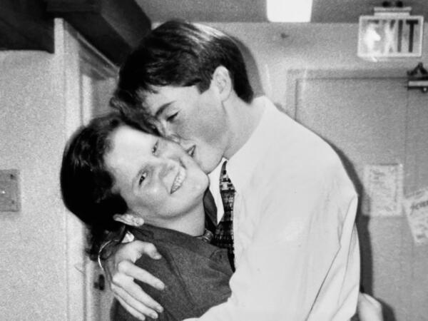 A man and woman hugging, man is kissing the woman on the cheek as she smiles at the camera