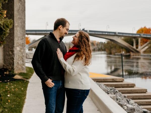 A couple standing outside at Trent campus, looking at each other lovingly, with bridge in the background