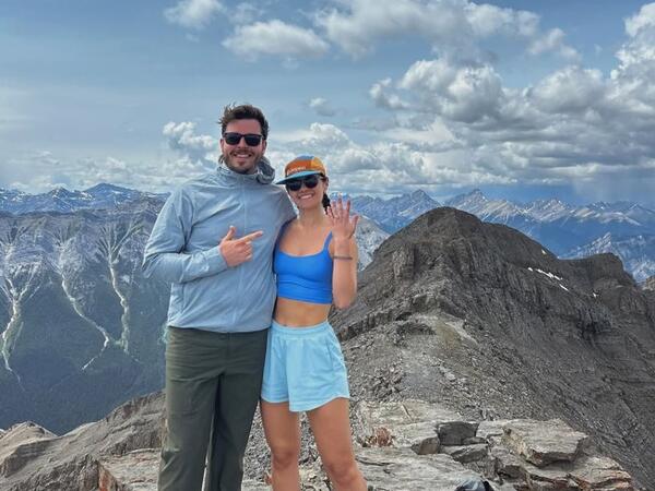 A couple standing on top of a mountain with the woman holding up her ring finger to announce engagement