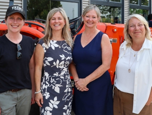 Dr. Karen Thompson, Tamara Lightbody, Deb Keay, and Dr. Cathy Bruce stand smiling in front of an orange tractor at the Trent Farm Research Centre. 