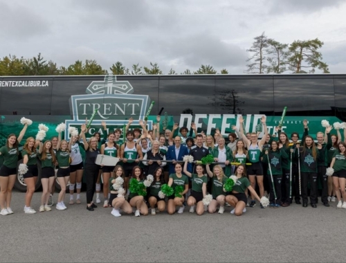 A large group of student athletes and Trent staff cheer in front of the new bus.