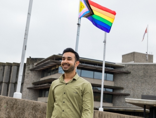 Sam Sarin smiles on campus with a Pride flag behind him