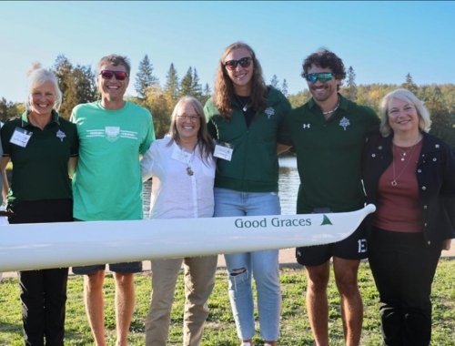 A group of people including Trent president Cathy Bruce and alumni stand in front of the new rowing scull.