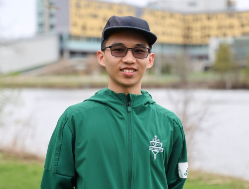 A man with a black ballcap standing outside of Gzowski College with the river in the background