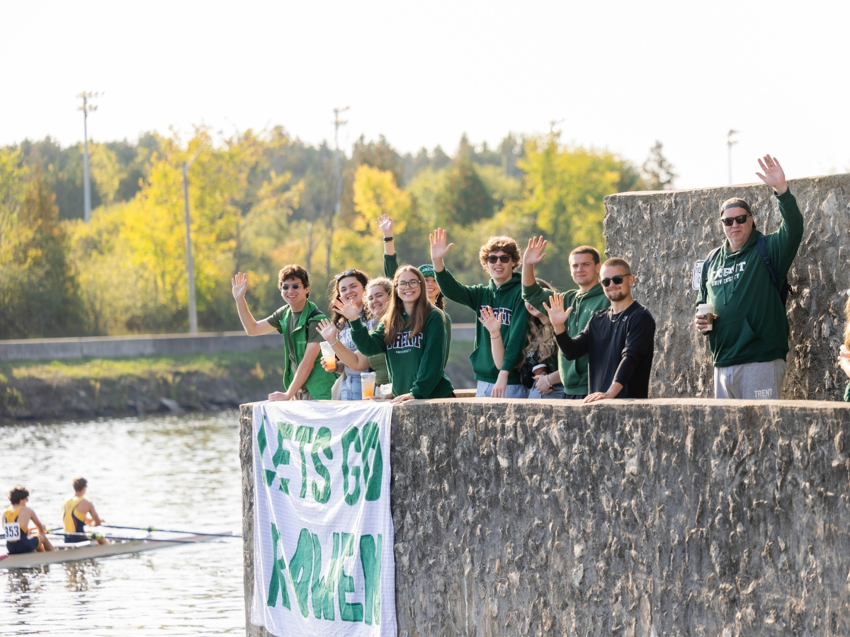 People standing beside the river, waving at the camera, with rowers in the water
