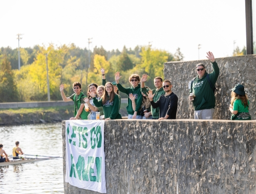 People standing at the water in Trent gear, with a large sign, cheering on rowers