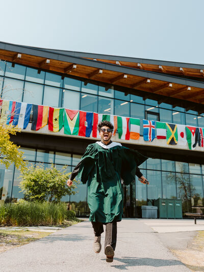 Young man in convocation regalia running at the camera with a wall of international flags behind