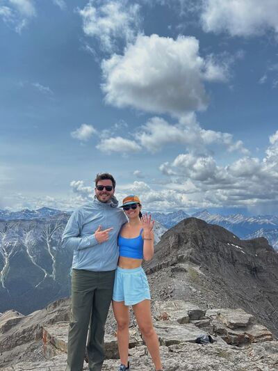 A couple standing on top of a mountain with the woman holding up her ring finger to announce engagement