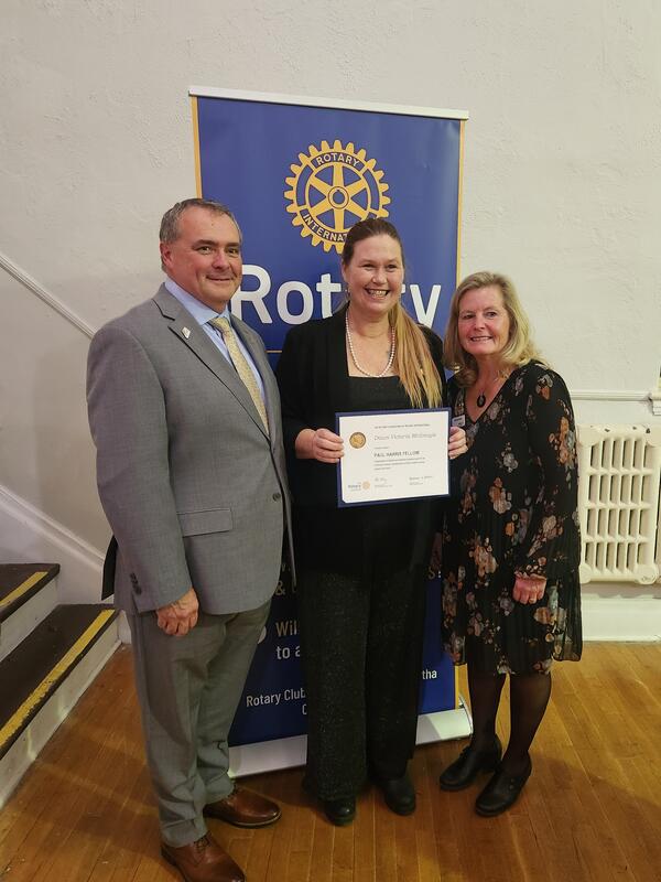 Three people standing infront of a Rotary sign, one is holding a certificate