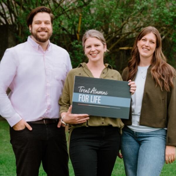 Three people smile holding a sign stating Alumni For Life